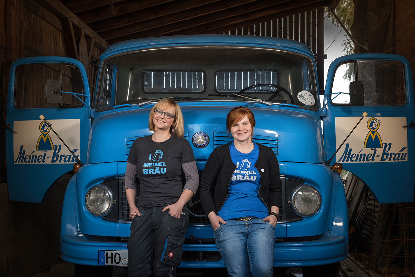 Zwei junge Frauen mit Meinel-Bräu-Shirts stehen vor der Front eines blauen Oldtimer-Lasters mit geöffneten Türen.