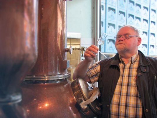 Ein älterer Herr im karierten Hemd steht an einer kupfernen Brennblase. Er hält ein Stielglas hoch und betrachtet den klaren Inhalt.